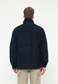 Άνδρας φορώντας ένα σκούρο μπλε φλις σακάκι με ψηλό γιακά και μπεζ cargo παντελόνι, εμφανιζόμενος από πίσω σε άσπρο φόντο.