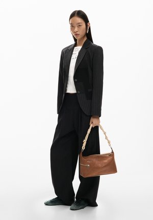 Kvinde iført stort sort jakkesæt med nålestriber og hvid top, der holder en brun læderhåndtaske med knyttet rem, stående foran hvid baggrund.