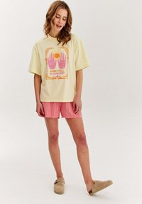 Krémové tričko s růžovou grafikou rukou a slunce, spárované s červenými pruhovanými šortkami a béžovými slip-on botami, vystavené na neutrálním pozadí.