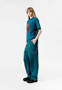 Camiseta de gran tamaño en color verde azulado con estampado gráfico, combinada con pantalones de chándal a juego en verde azulado que presentan un detalle de cadena lateral. Zapatillas negras de textura y estilo slip-on.