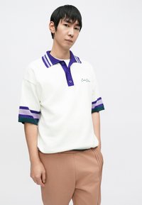 Biała koszulka polo z krótkim rękawem, z fioletowym kołnierzem i paskami w kolorach fioletowym i zielonym. Posiada zapięcie na guziki oraz haftowane logo.