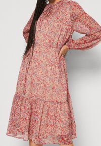 Robe midi florale en tissu léger et transparent, présentant un motif rose et rouge, des manches longues et un ourlet à volants en cascades. Liens au cou.