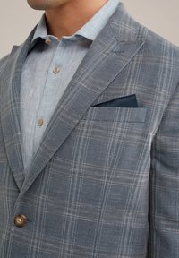 Hellblaues Leinenhemd unter einem grauen karierten Blazer mit braunen Knöpfen und einer marineblauen Einstecktuch. Strukturiertes Gewebe mit klaren Linien und Mustern.