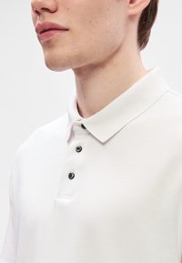 Bílá polo košile s klasickým límečkem, třemi černými knoflíky a hladkým textilním povrchem. Detailní pohled, viditelné rameno a krk.