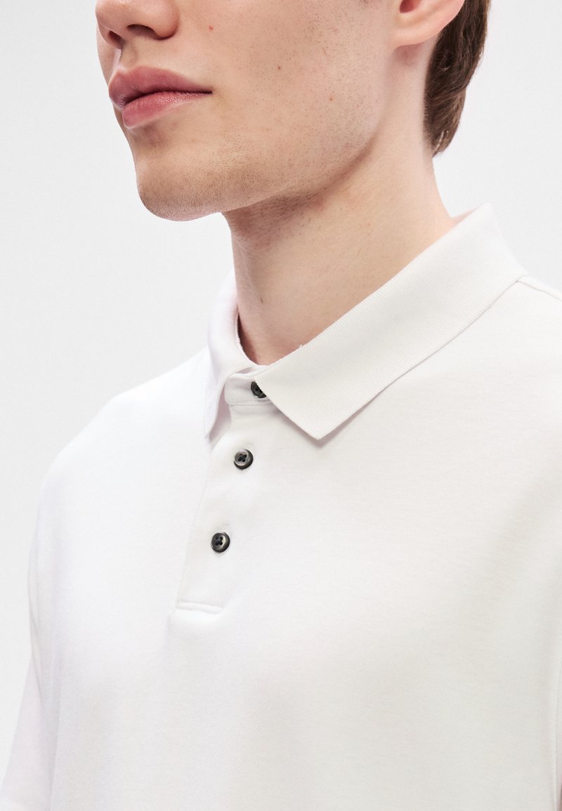 Polo blanc avec un col classique, trois boutons noirs et une texture de tissu lisse. Vue en gros plan, épaule et cou visibles.