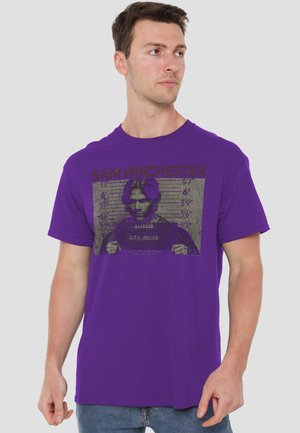 Man wearing a purple T-shirt with a black graphic mugshot of Sam Winchester and height chart in the background.