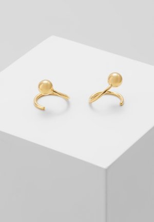 Guld åbne ringe med hver en glat rund kugle på toppen, udstillet på en hvid kubisk piedestal.
