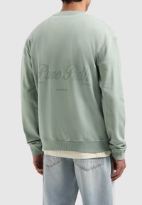 Mint green sweatshirt with ribbed cuffs and hem, featuring embossed "Pure Path" text on the back. Soft fabric. Worn over a light shirt.
