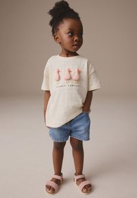 Lys beige skjorte med lyserøde fugleapplikationer og teksten "Always Curious", kombineret med blå denimshorts og lyserøde sandaler. Simpelt ternet mønster.