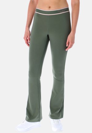 Femme portant des leggings évasés taille haute vert olive avec une ceinture bordée de rose portant la mention Ted Baker Sport et des baskets blanches.
