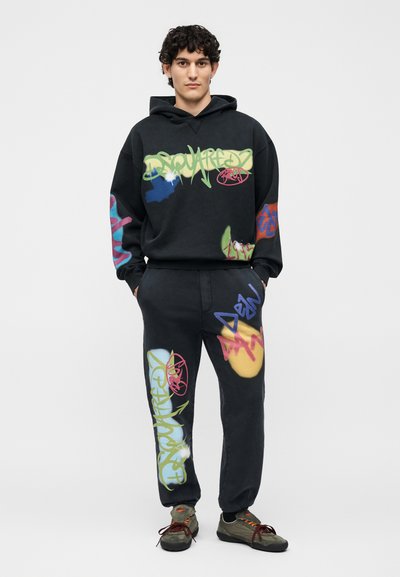 Νέος άνδρας με σγουρά μαλλιά που φοράει μαύρο φούτερ με κουκούλα και φόρμες με πολύχρωμα σχέδια σε στυλ γκράφιτι, στέκεται με τα χέρια στις τσέπες.