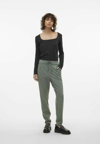 Zwarte langemouwen top met een vierkante halslijn, gecombineerd met losse, salie groene broek met een trekkoord in de taille. Draag met zwarte plateau schoenen.