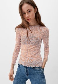 Langärmliges Shirt aus transparenter Stoff, mit einem wirbelnden Muster in zartem Orange und Weiß. Mit geschwungenem Saum und strukturierter Oberfläche. Kombiniert mit blauen Jeans.