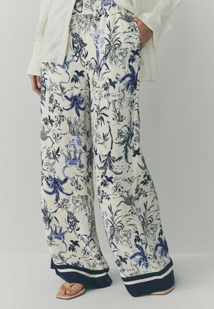 Wijdvallende crèmekleurige broek met navy- en lavendelkleurige dier- en plantenprints, gedragen met beige sandalen en een wit shirt met lange mouwen.