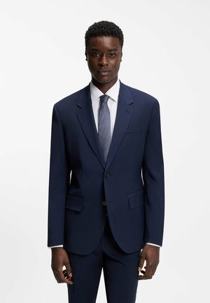 Homme portant un costume bleu foncé, une chemise blanche et une cravate bleue, debout devant un fond blanc uni, fait face à l’avant.