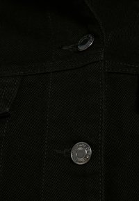 Svart denimtyg med en silverknapp och subtila sömnadsdetaljer. Texturen är grov med ett svagt vertikalt mönster.