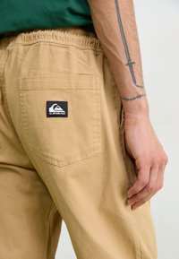 Tan cargo bukser med elastisk talje, med en baglomme med et sort og hvidt Quiksilver-logo. Glat stoffarve.