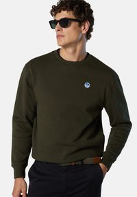 Sweatshirt vert olive avec col ras du cou, poignets côtelés et logo sur la poitrine. Associé à un pantalon sombre et une ceinture texturée avec un accent marron.