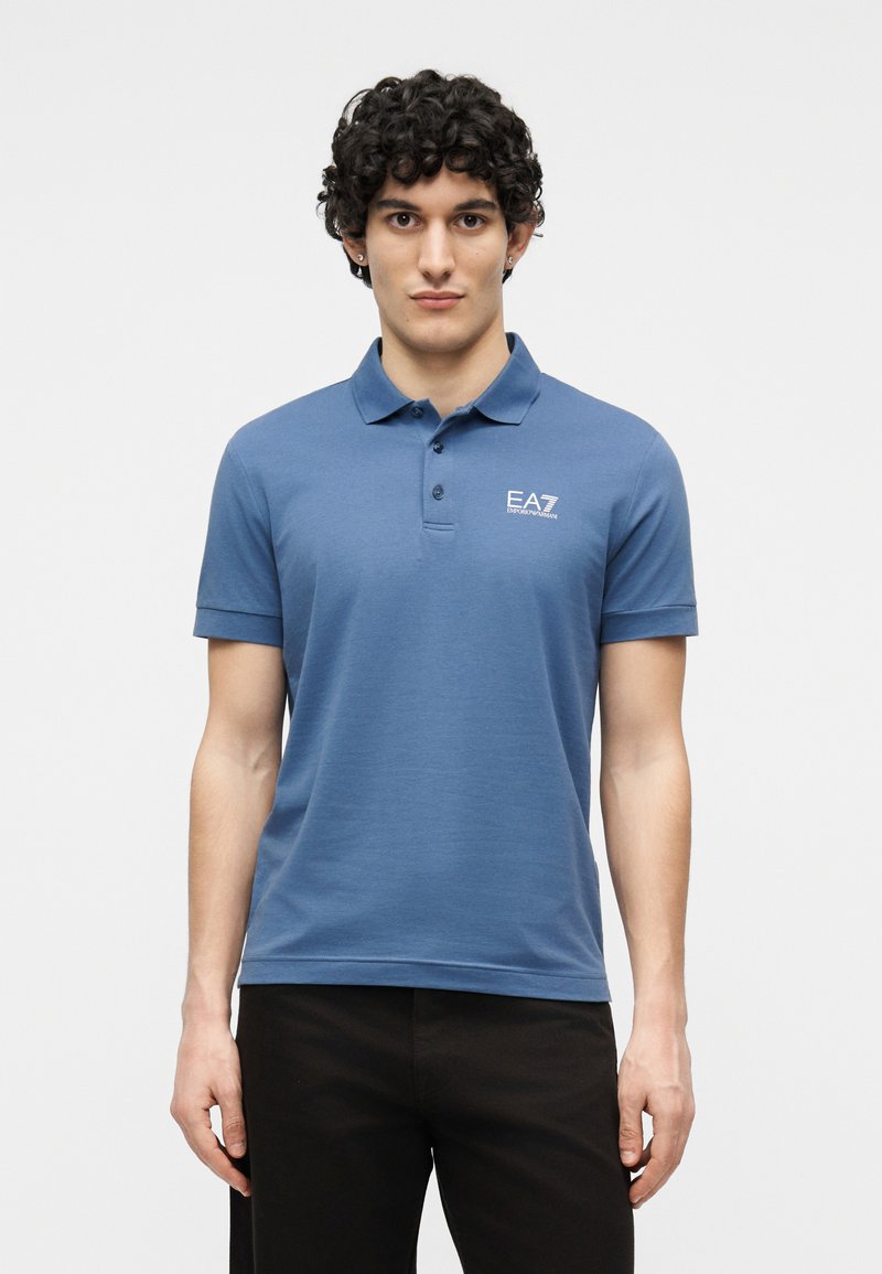 Νεαρός άνδρας με κατσαρά μαλλιά, φορώντας μπλε πόλο μπλούζα EA7 και μαύρο παντελόνι, στέκεται μπροστά σε απλό ανοιχτό φόντο.