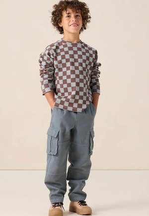 Jongen met krullend haar, in een bruin en lichtblauw geruit sweater, grijze cargo broek en bruine schoenen, staand met zijn handen in zijn zakken.