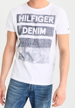 Camiseta blanca de algodón con un estampado gráfico en gris que dice "HILFIGER DENIM", con mangas cortas y cuello redondo.