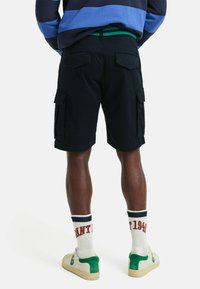 Pantalones cortos cargo negros con bolsillos laterales, una cintura elástica verde y bolsillos traseros. Calcetines blancos con texto rojo y azul, combinados con zapatillas con detalles verdes.