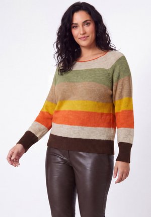 Vrouw met donker krullend haar, gekleed in een meerkleurige gestreepte trui en donkerbruine leren broek, staand tegen een witte achtergrond.
