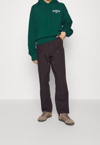 Sudadera con capucha verde con logo blanco, combinada con pantalones marrón oscuro que presentan remaches dorados. Zapatillas marrones con rayas completan el conjunto.