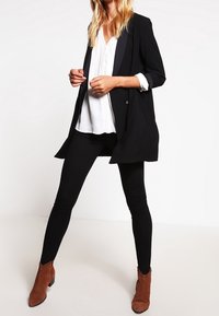 Černý blazer, bílá blůza a černé legíny; hnědé kotníkové boty. Blazer má mírně oversized střih a saténový klopový detail.