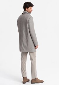 Abrigo de lana gris con un cuello pronunciado, diseño de longitud media y detalles en las costuras laterales, combinado con pantalones claros y botas de tobillo marrones.