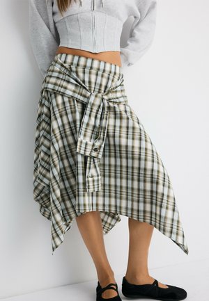 Femme portant une jupe asymétrique à carreaux verts et blancs et un sweat à capuche court gris clair, avec des chaussures noires sans lacets, sur fond blanc.