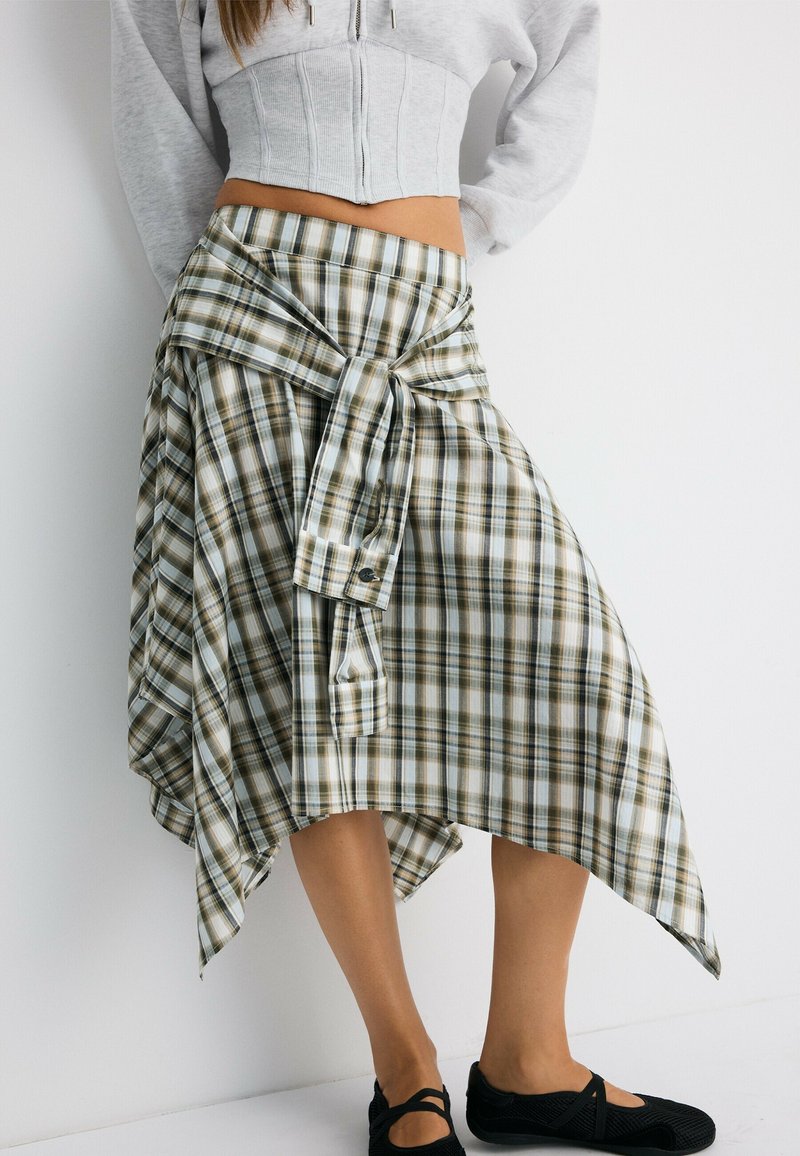 Femme portant une jupe asymétrique à carreaux verts et blancs et un sweat à capuche court gris clair, avec des chaussures noires sans lacets, sur fond blanc.