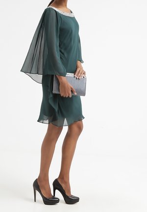 Szmaragdowozielona sukienka z szyfonu z przezroczystymi rękawami i zdobionym kamieniami dekoltem, zestawiona ze srebrną kopertówką i czarnymi szpilkami.