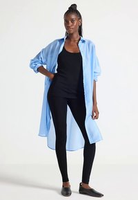 Chemise bleue claire à boutons, manches longues, tissu semi-transparent, coupe décontractée ; accompagnée d'un débardeur noir et de leggings noirs.
