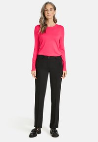 Langärmliges, leuchtend pinkes Oberteil und eng anliegende schwarze Hose mit glatter Textur. Schwarze Schuhe mit dicker Sohle. Neutraler Hintergrund.