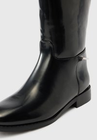 Λείο μαύρο δερμάτινο μπότες μέχρι το μέσο της γάμπας με χαμηλό τακούνι και απλό λουράκι στο πλάι, πάνω σε απλό φωτεινό φόντο.