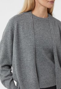 Szary sweter na drutach z dekoltem w łódkę, otwarty z przodu, z prążkowanym dołem. Miękka tekstura, luźny krój i delikatne cętki na całej tkaninie.