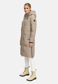 Kvinna med en beige lång pufferjacka med huva, vita uppvikta byxor och beige snörstövlar mot en enkel bakgrund.