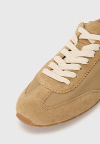 Tan suede sneaker with beige accents, featuring a rounded toe, prominent laces, and stitching details on the upper.