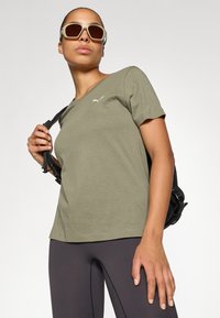 Camiseta de manga corta verde oliva con un sutil logo blanco, combinada con leggings negros y una mochila negra, llevada con gafas de sol.