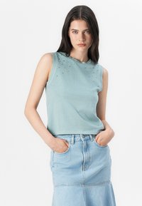 Modellen har på seg en ermeløs lyseblå topp med dekorative detaljer, sammen med et lyst denimskjørt, og står foran en ensfarget bakgrunn.
