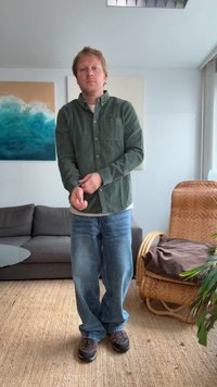 Camicia verde a maniche lunghe, strato chiaro sotto, jeans blu e scarpe scure. Stanza con divano grigio, pianta e sedia intrecciata.