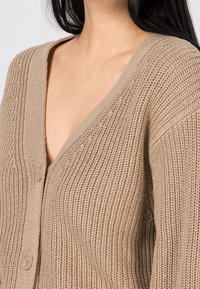 Beżowy sweter z dzianiny z dekoltem w kształcie V i teksturowanym materiałem, z dużymi guzikami z przodu i żeberkowymi wykończeniami na brzegach.