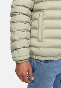 Lys grøn pufferjakke med vandret quiltet design, elastiske manchetter og en sidelomme, parret med lyseblå jeans.