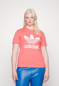 Koralā rozā T-krekls ar baltu Adidas logo un trīs svītru dizainu, īsām piedurknēm un klasisku apaļu kakla izgriezumu, valkāts kopā ar ziliem sp�īdīgiem biksēm.