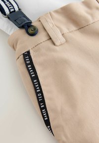 Primo piano di pantaloni beige con banda laterale nera etichettata "Baker by Ted Baker" e bretelle blu navy con asola per bottone.