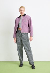 Paarse cropped jack met zwarte kraag, grijs aansluitend topje, geprinte olijfgroene broek en zwarte schoenen op een lichtgroene vloer.