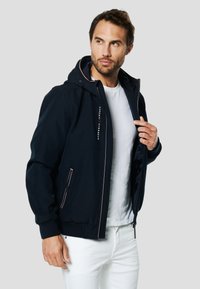 Blauer Zip-up Pullover mit Kapuze, seitlichen Taschen, gerippten Bündchen und einem Logo. Trägt man über einem weißen T-Shirt und kombiniert mit weißen Hosen.
