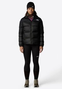 Jauna sieviete melnā pūkainā jaku, melnās legingās, bēša pārgājienu apavos, melnā cepurē un rakstainā kakla lakatā, stāvot pret balto fonu.