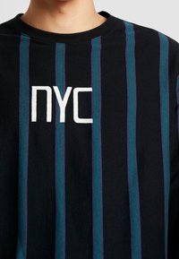 Svart bomullströja med vertikala blå och gröna ränder, med vit "NYC"-text broderad i mitten. Rund halsringning, avslappnad passform.